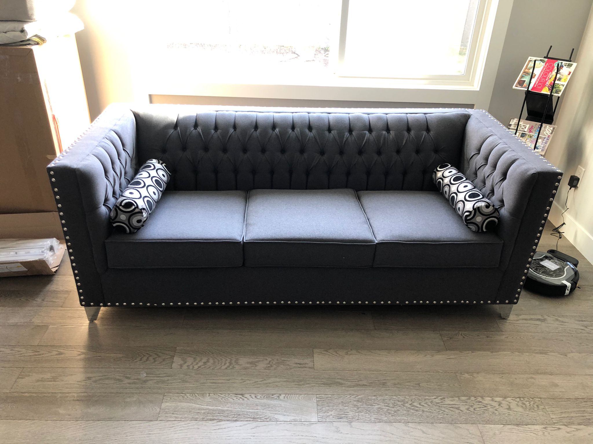 Gray tufted sofa with patterned bolster pillows on wooden floor, placed near a window