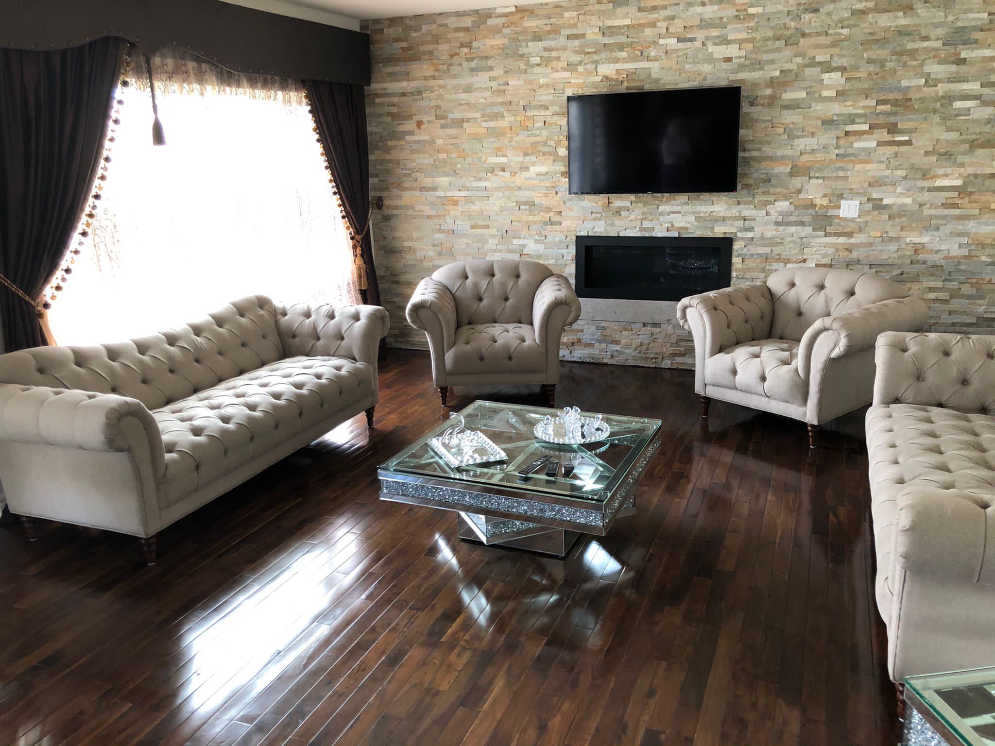 Elegant living room with beige tufted sofas and armchairs, glass coffee table, wooden floor, stone accent wall, and wall-mounted TV.