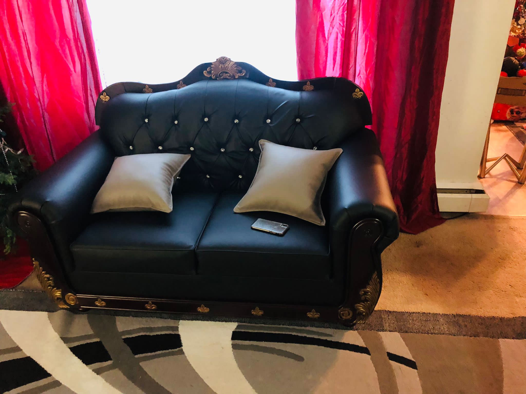 Black leather tufted loveseat with two beige pillows in front of red curtains.
