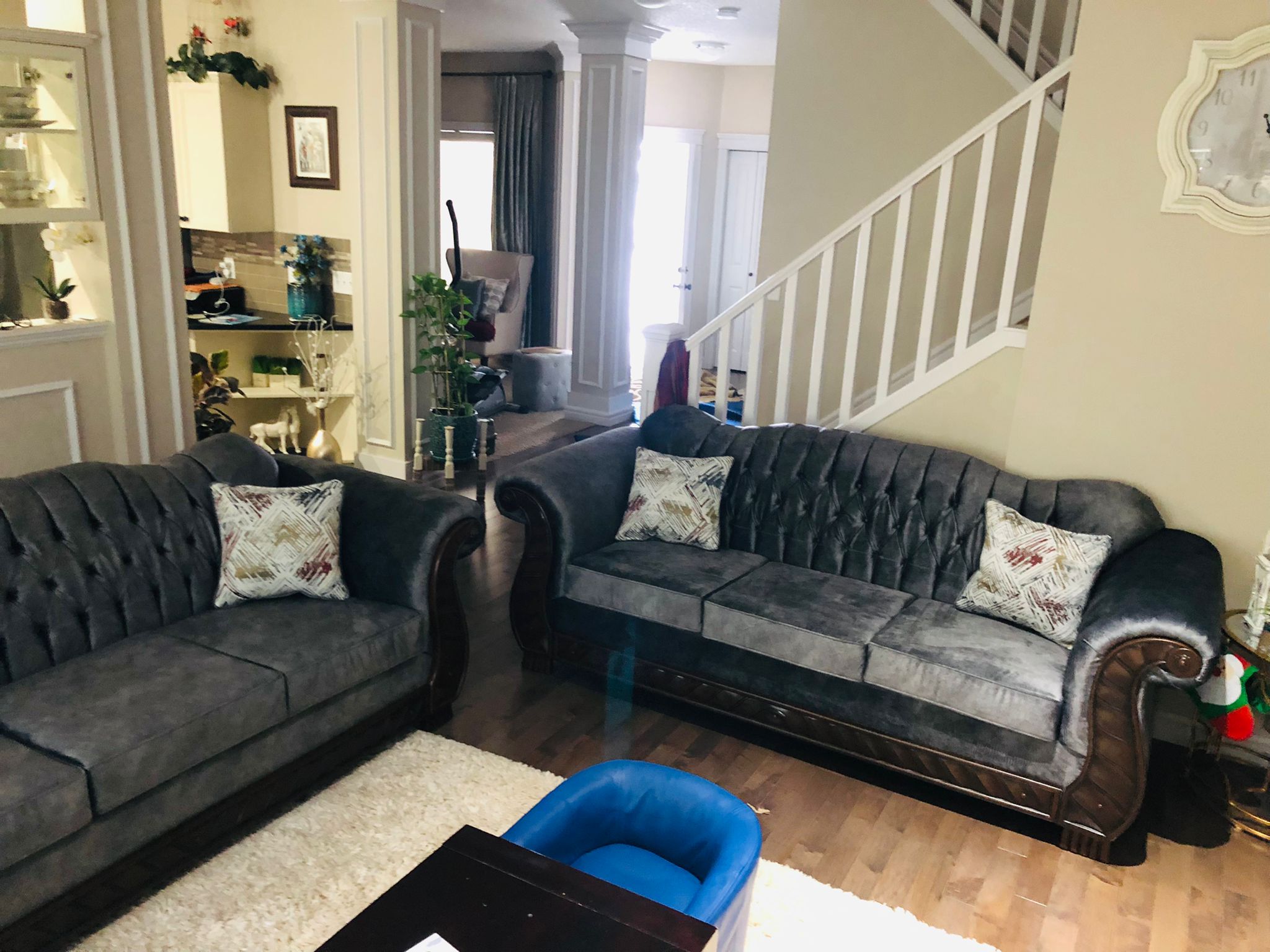 Two gray tufted sofas with patterned pillows in a living room with wooden floor and a blue seat.