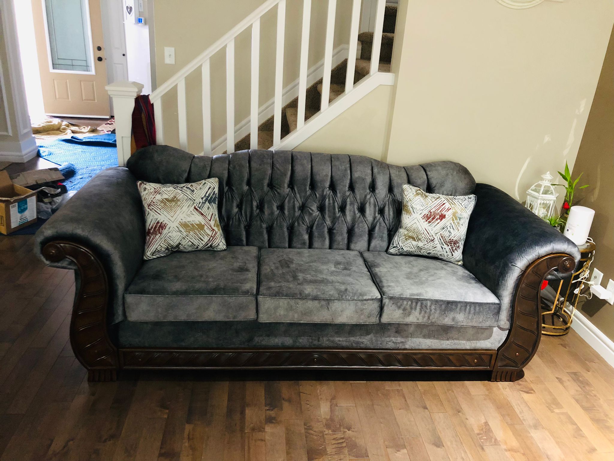 Gray tufted sofa with wooden trim and two patterned pillows placed near a staircase in a living room.